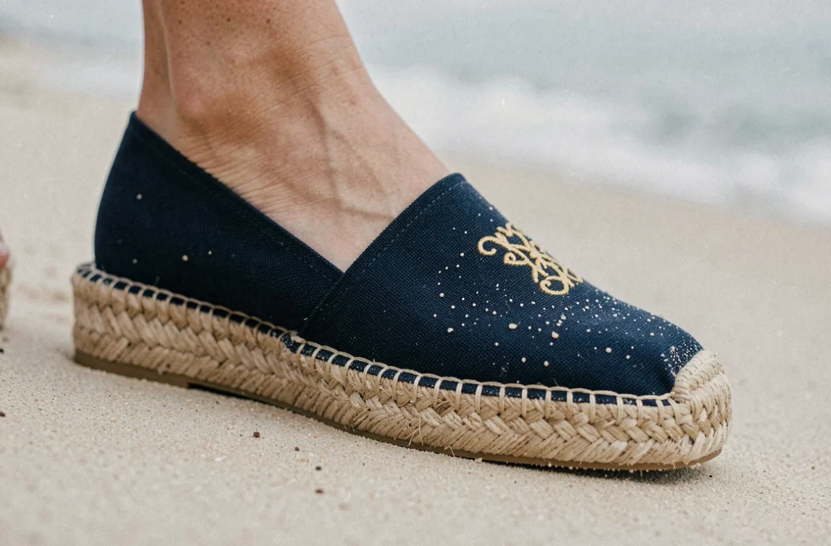 Détail rapproché d'une espadrille de luxe en toile marine avec monogramme doré et semelle en jute tressée sur sable de plage